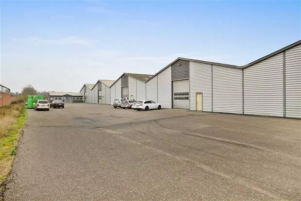 Lagerhal med flere parkerede biler og gårdhave med græs. Garageporten er åben og der er en stor parkeringsplads