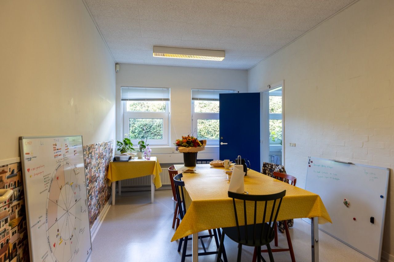 Lyse lokaler med to borde, gule duge og lyserøde blomster i en vase. To skriveborde med en hvid væg i baggrunden.