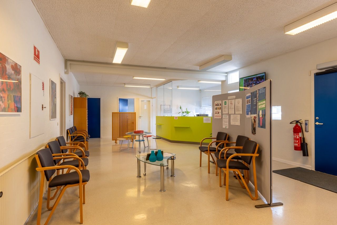 En lyse lobby med moderne møbler og en gulvlampe. En lysegul væg med et skriftligt meddelelse. Et rødt ildslukker er