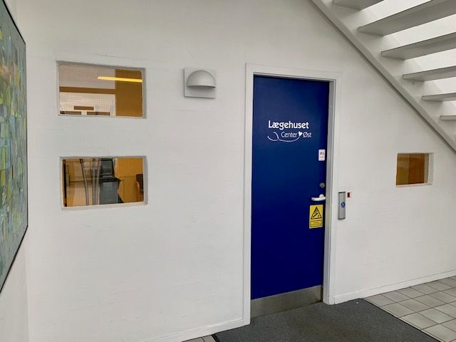 En hvid væg med to vinduer, der er åbne og stiger op til trapper. Der er en blå dør med teksten Laegehuset Center Ost.