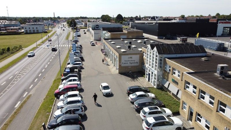 Luftfoto af Sundhedshuset Love Apotek med mange parkerede biler og et menneske, der går gennem parkeringspladsen