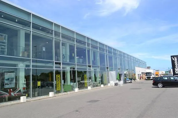 En stor butiksbygning med store vinduer, der reflekterer himlen og luften. Foran er parkeringspladsen med en række hvide