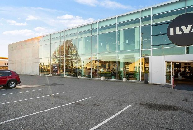 Glass facade med logoet ILVA og indgangen. Et rødt køretøj parkeret ved siden af. En plante i en pot og en søjle med