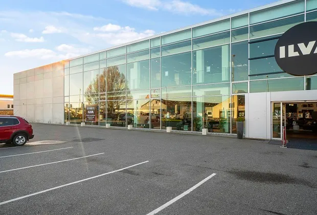 Glass facade med logoet ILVA og indgangen. Et rødt køretøj parkeret ved siden af. En plante i en pot og en søjle med