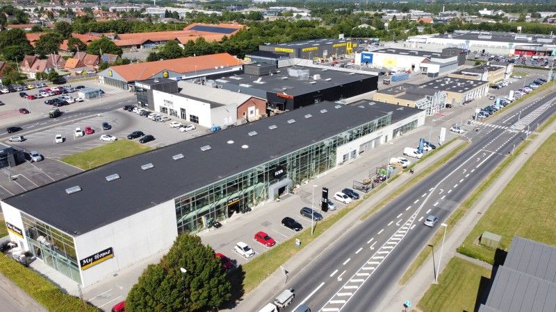 Her ses et stort forretningscenter med flere biler parkeret. Der er butikker, et stort parkeringsområde, en hovedvej og