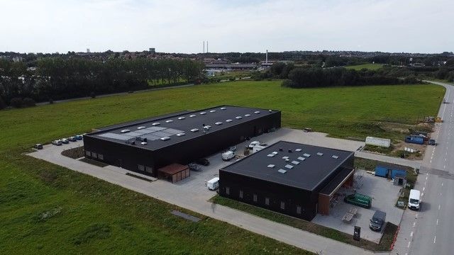 To moderne bygninger med solceller på tag og parkeringsplads, omgivet af grønt landskab og en vej med biler og busser i
