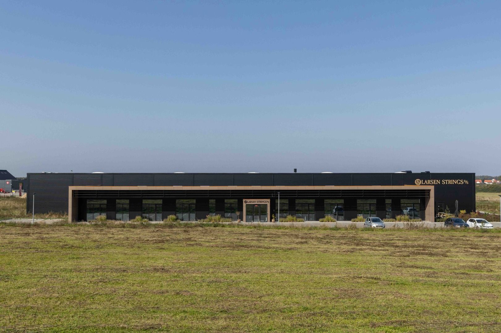 Larsen Strings logo er anbragt på bygningens facade med to biler parkeret foran og en græsplæne i baggrunden