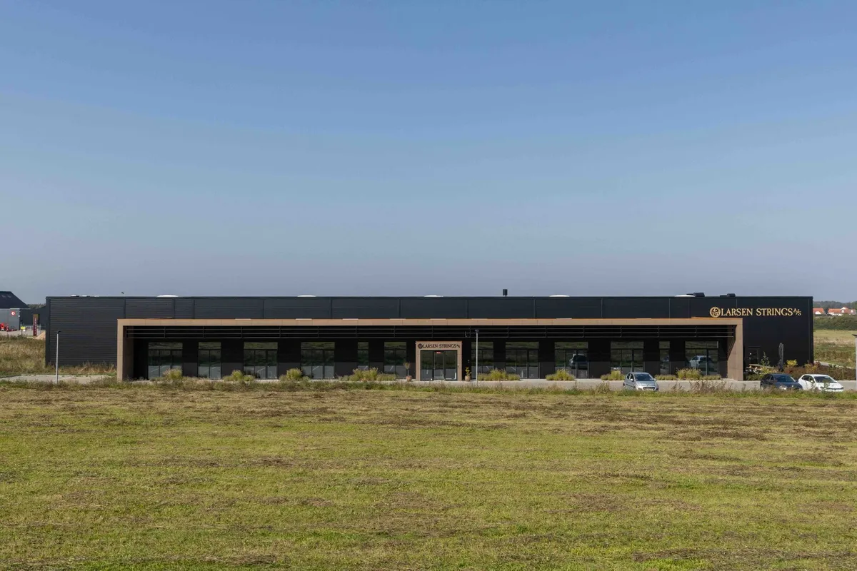 Larsen Strings logo er anbragt på bygningens facade med to biler parkeret foran og en græsplæne i baggrunden