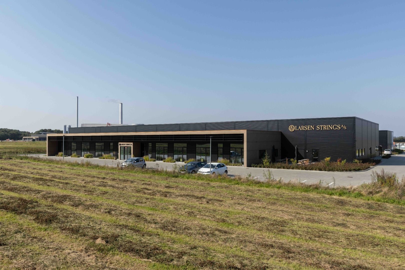 Larsen Strings produktionsbygning i Holstebro med tre parkerede biler og to høje skorstene i baggrunden