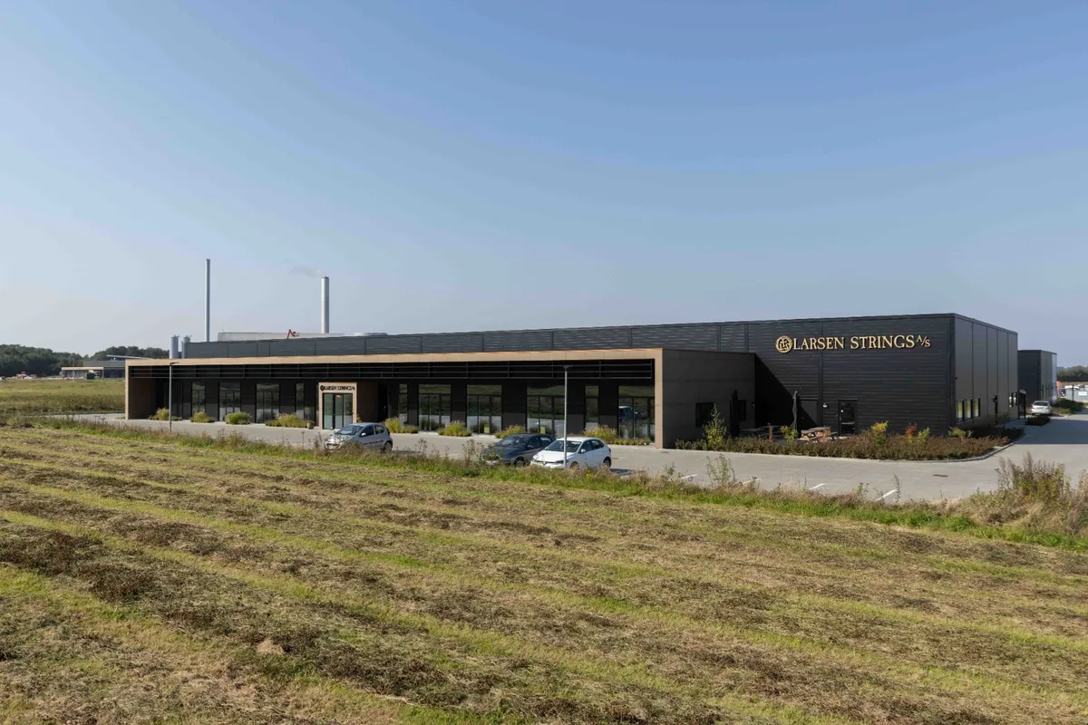 Larsen Strings produktionsbygning i Holstebro med tre parkerede biler og to høje skorstene i baggrunden