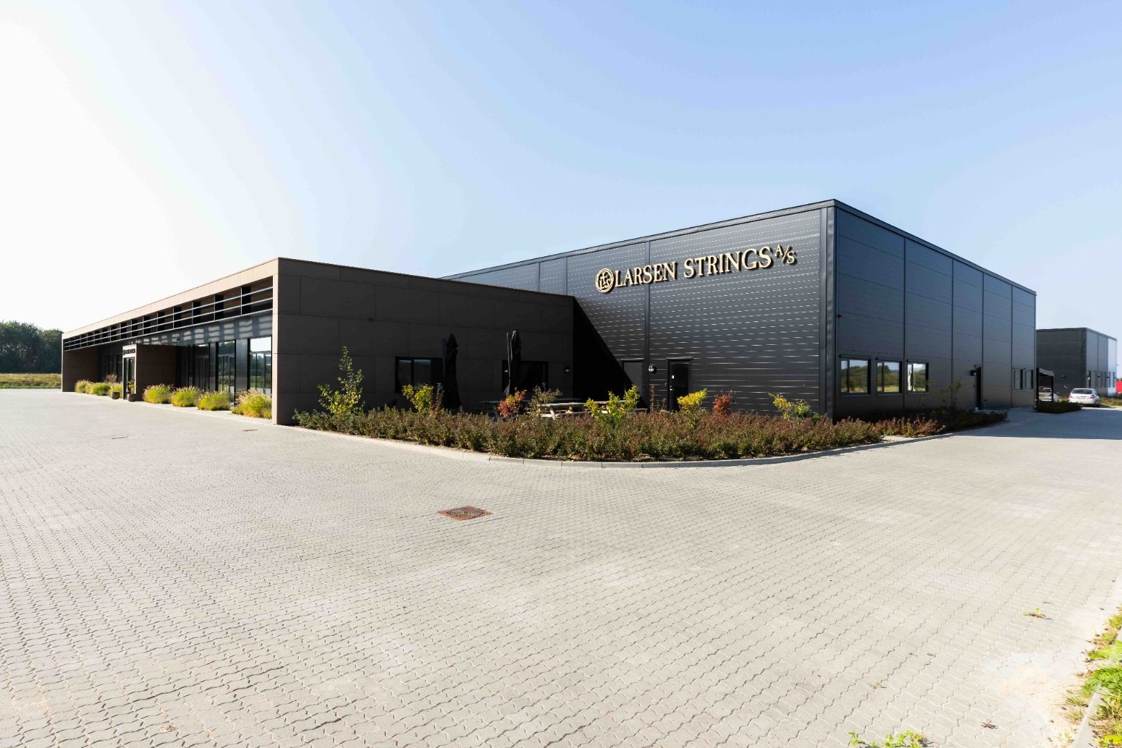 Larsen Strings logo præsenteret på en stor bygning med flere indgange og terrasser med parasoller. Foran bygningen er der et