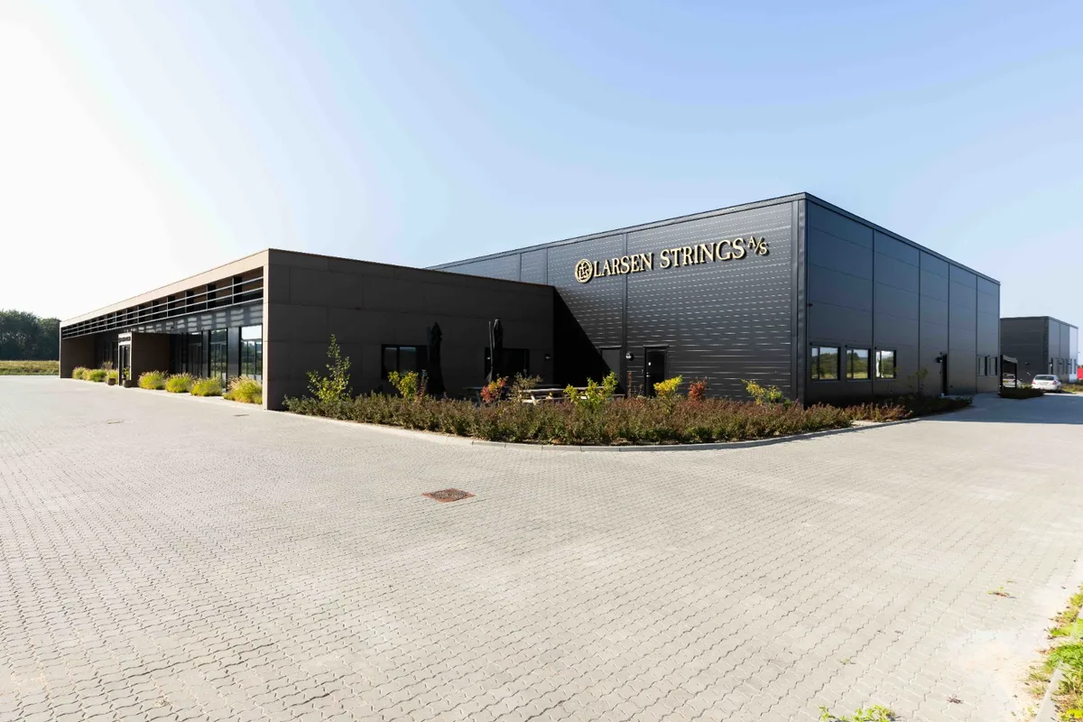 Larsen Strings logo præsenteret på en stor bygning med flere indgange og terrasser med parasoller. Foran bygningen er der et