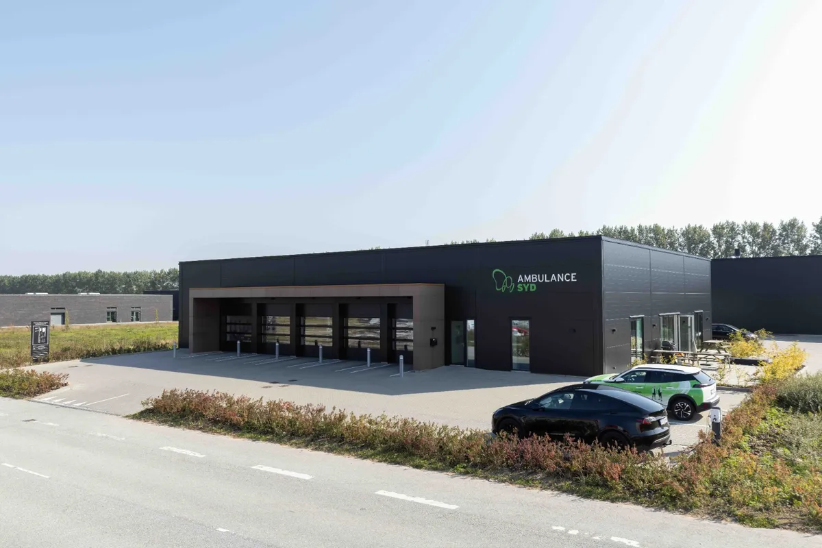 Ambulance Syd i bygningen med parkeringsplads og grønne planter med biler parkeret og en skov i baggrunden