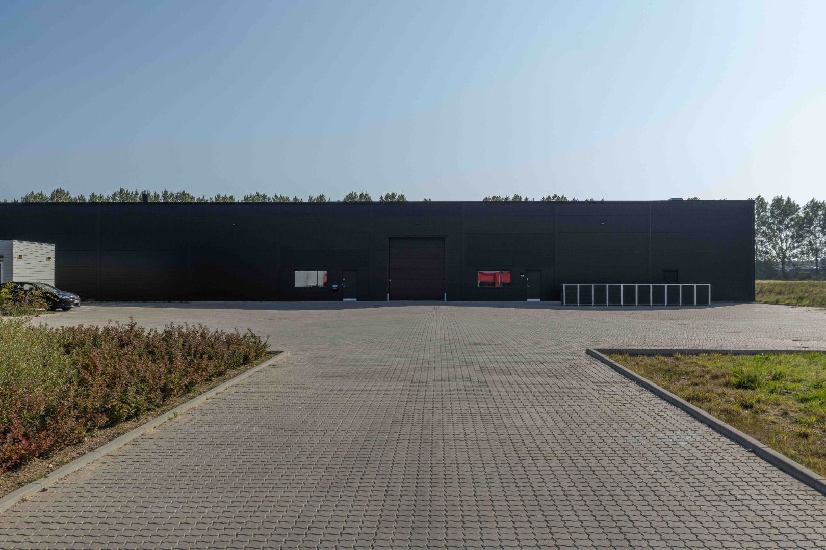 Sort bygning med dobbeltdør og lagerrum er placeret ved siden af en parkeringsplads og græsarealer med en bil parkeret i