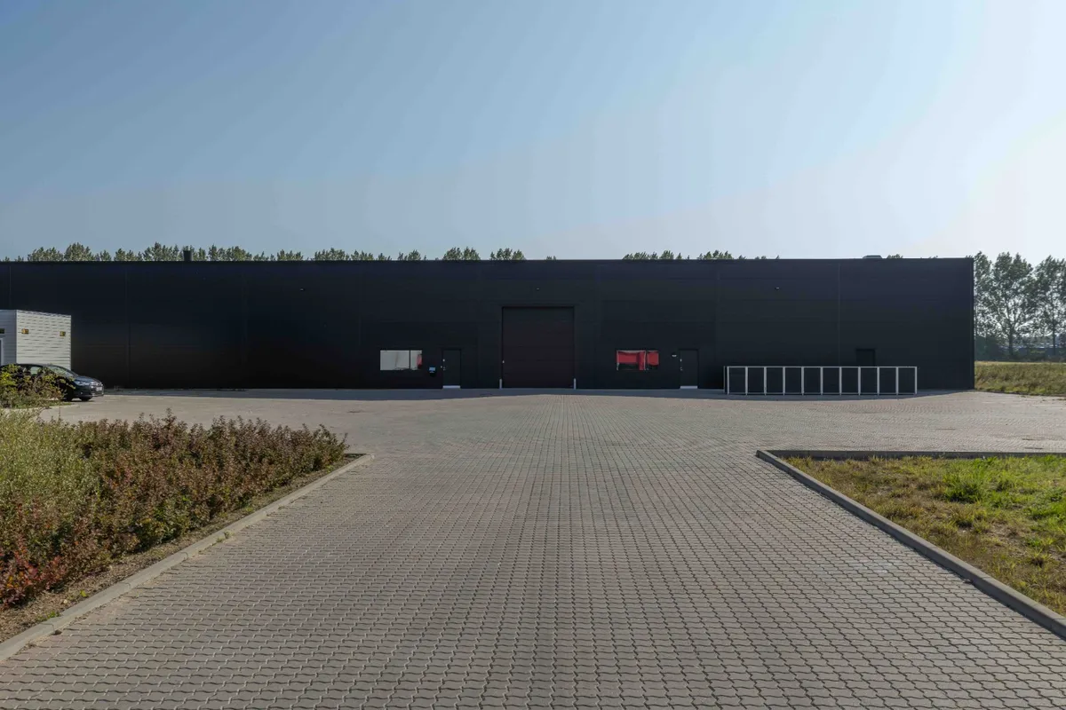 Sort bygning med dobbeltdør og lagerrum er placeret ved siden af en parkeringsplads og græsarealer med en bil parkeret i