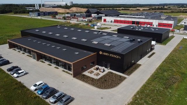 To bygninger med solpaneler på tagerne, parkeringsplads med biler og folk, grønne marker og bakker i baggrunden