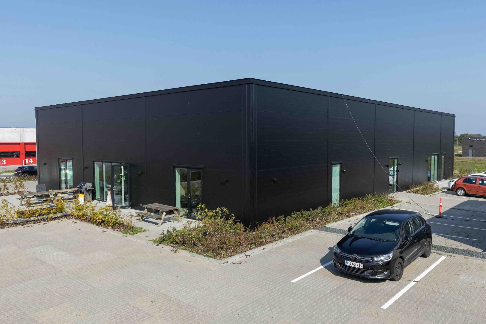 En nybygget sort bygning med glasruder og døre står på en parkeringsplads. Et bord med bænke står foran. Et