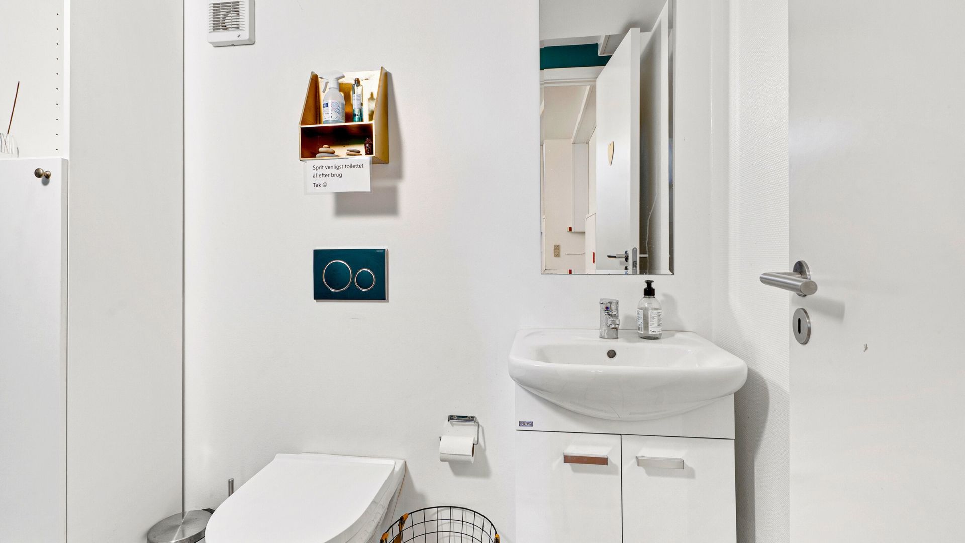 Hvidt toilet med moderne design, håndvask, to toiletpapirholdere, renlighedsprodukter og skrift med oplysning om toilettets