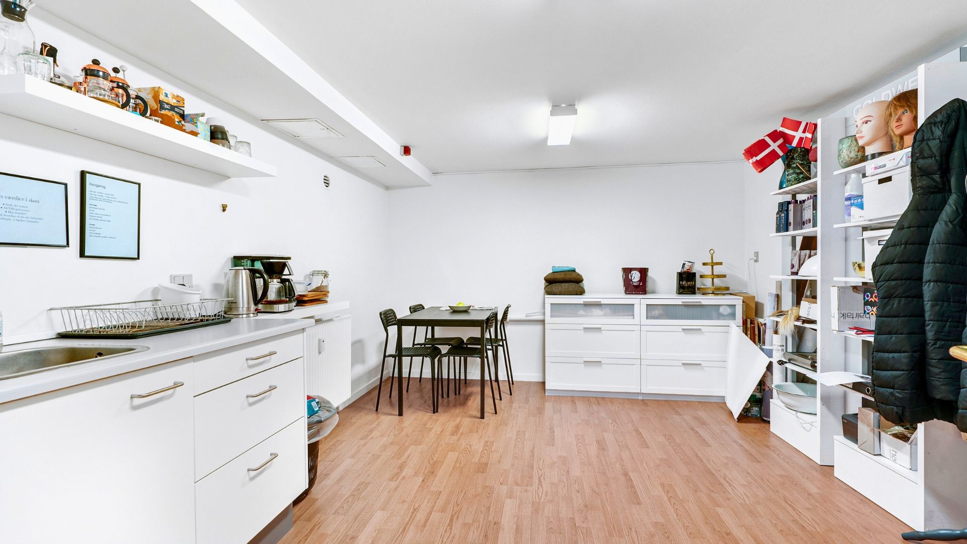 Minimalistisk køkken med hvide vægge, højloft, indbygget lyskabin, køkkenbord med stole, åbne hylder og et bord med bøger og