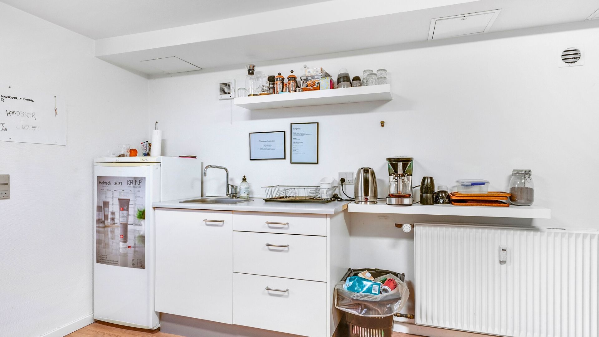 Et lille køkken med en køleskab, et skab, en radiator og et skrivebord med en skraldespand, en kaffemaskine og en