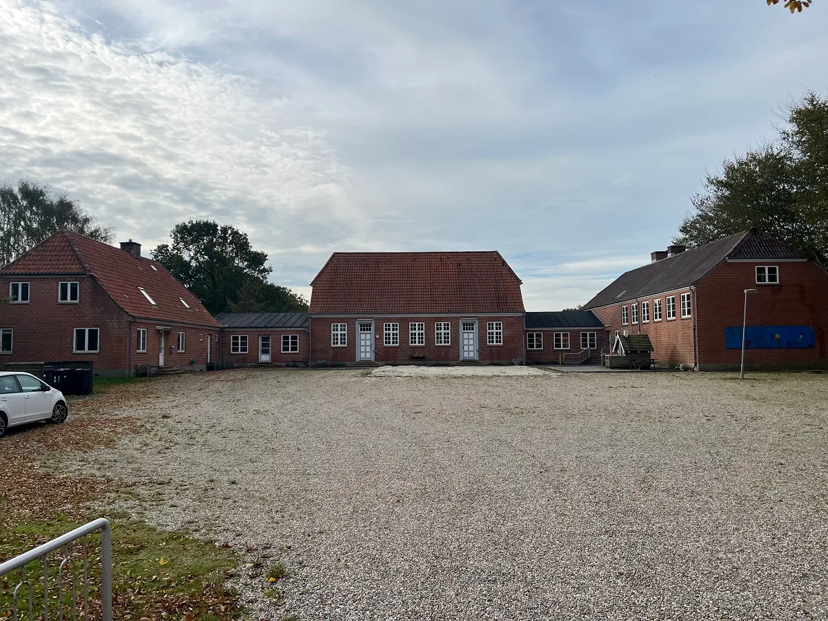 Fire røde bygninger med høje tag og hvide ruderuder og døre står på en grund med grus og græs.