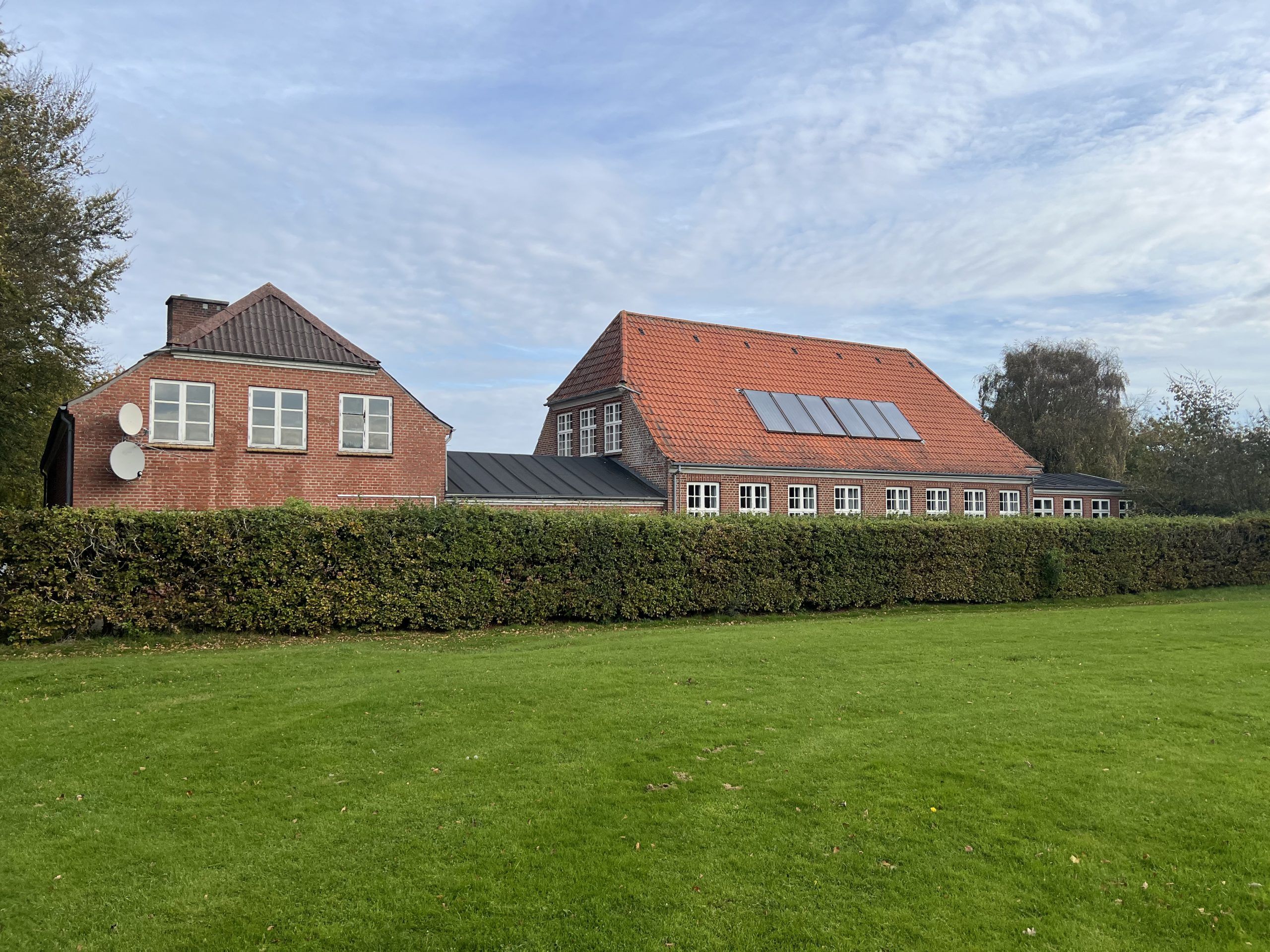 To store bygninger med røde tag og flere vinduer står bag en høj buskhæk og markeret med solpaneler