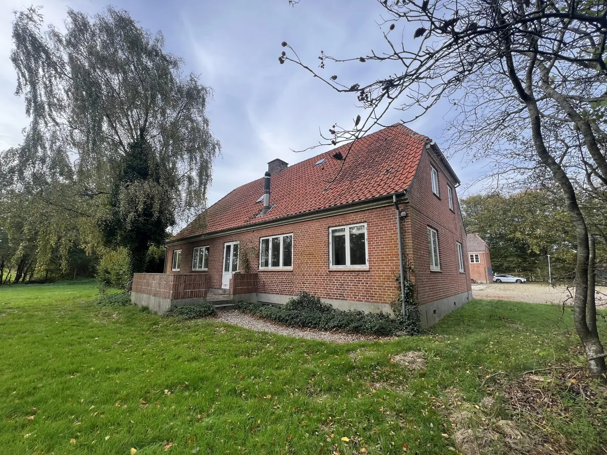 Et rødt tegltag på en bygning, der står midt i en grøn længde. Der er flere vindueruder og en række buske og træer omkring