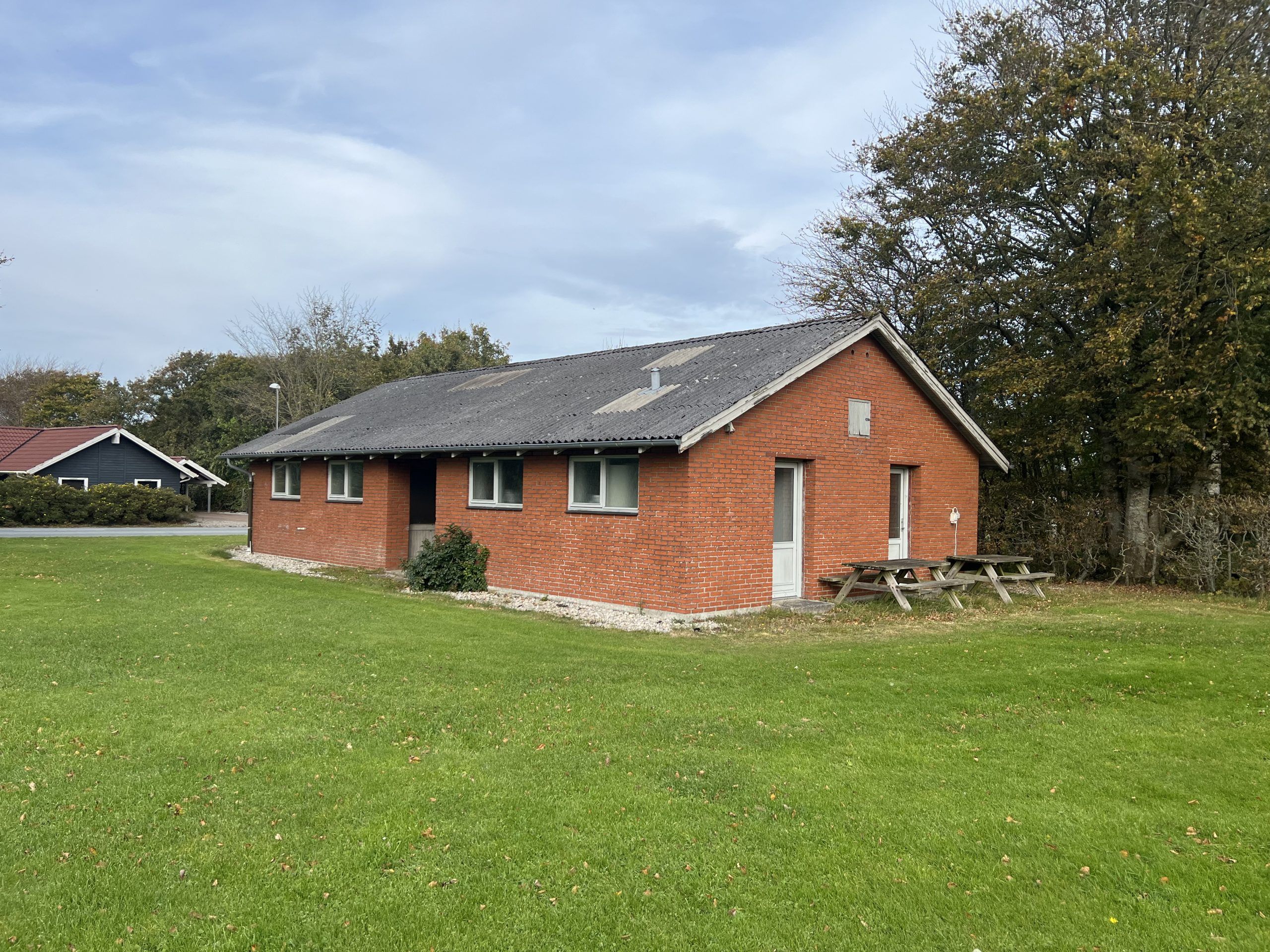En rødmalet sommerhusbygning med en tæt græsplæne og to bænke foran. En anden bygning med et rødmalet tag ses i baggrunden.
