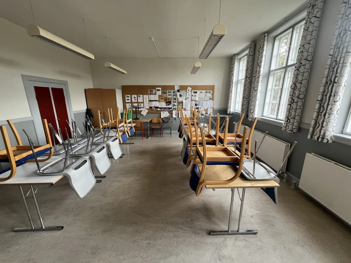 Klassenrum med to lange rækker af skolebænke, der er stillet op med stole, der vender mod væggen. Lampelys hænger fra loftet