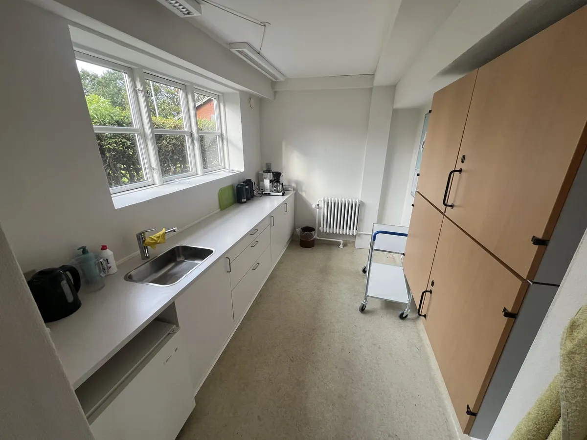 Køkken med hvide vægge, højloft og lys. Køkken med lyseskabe, radiator, skabe, og vindue med udsigt til træer og huse.