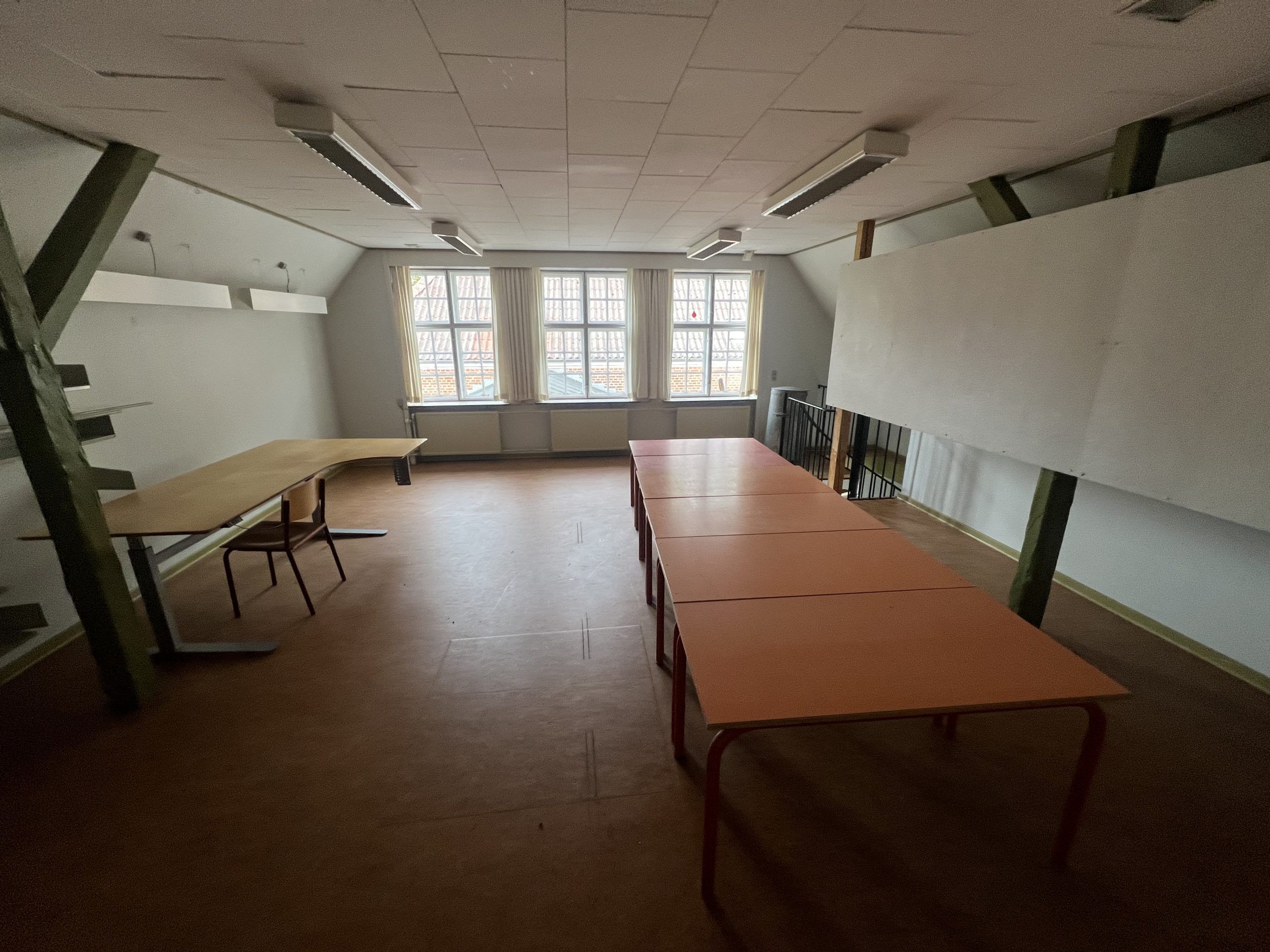 Et rum med lyse lofter, skråt liggende vægge og lange borde med stole samt lysekroner og vinduer