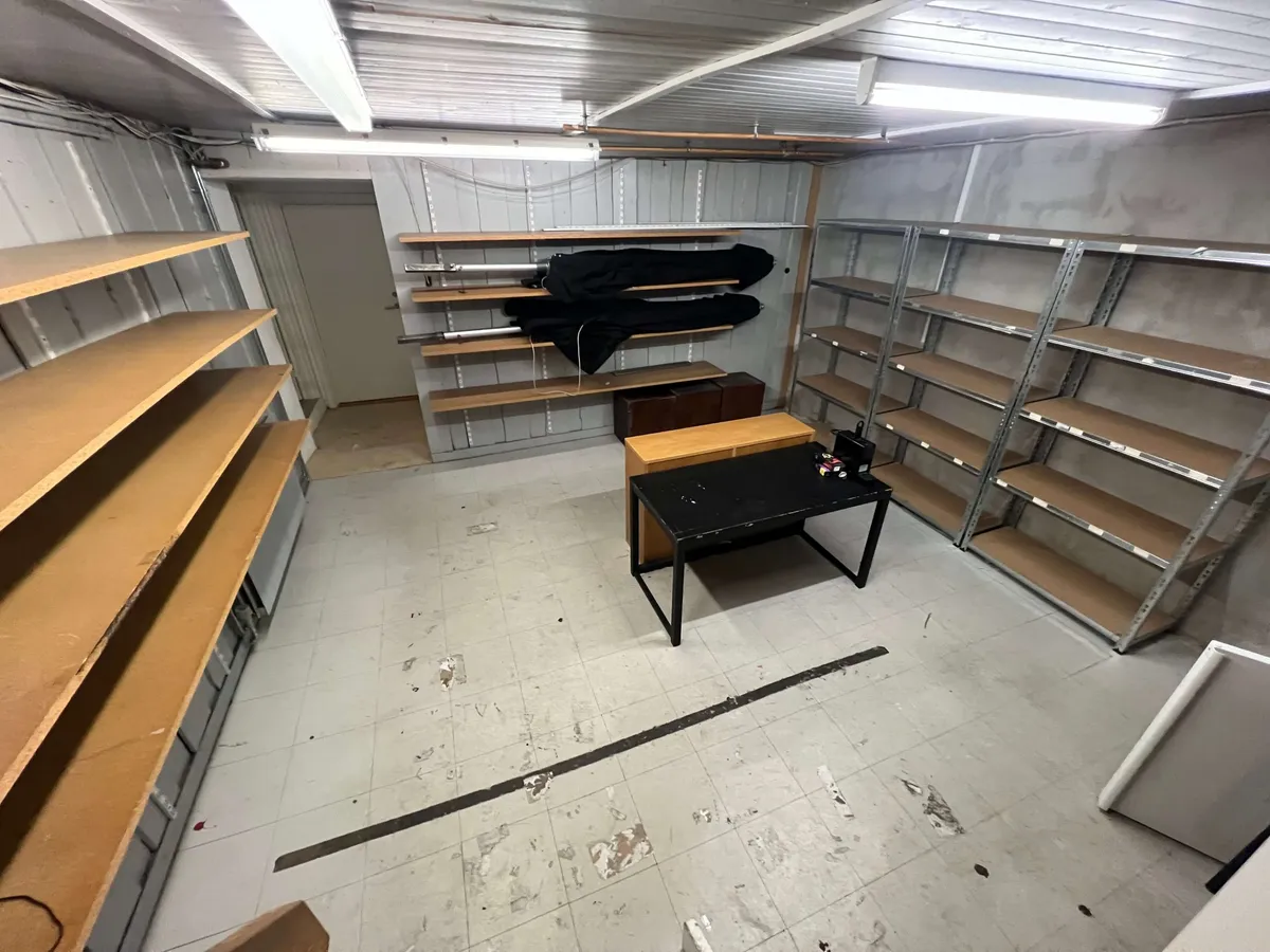 Et tomt lager med flere træhylder og en skrivebord samt en åben dør til et lille rum med tøj