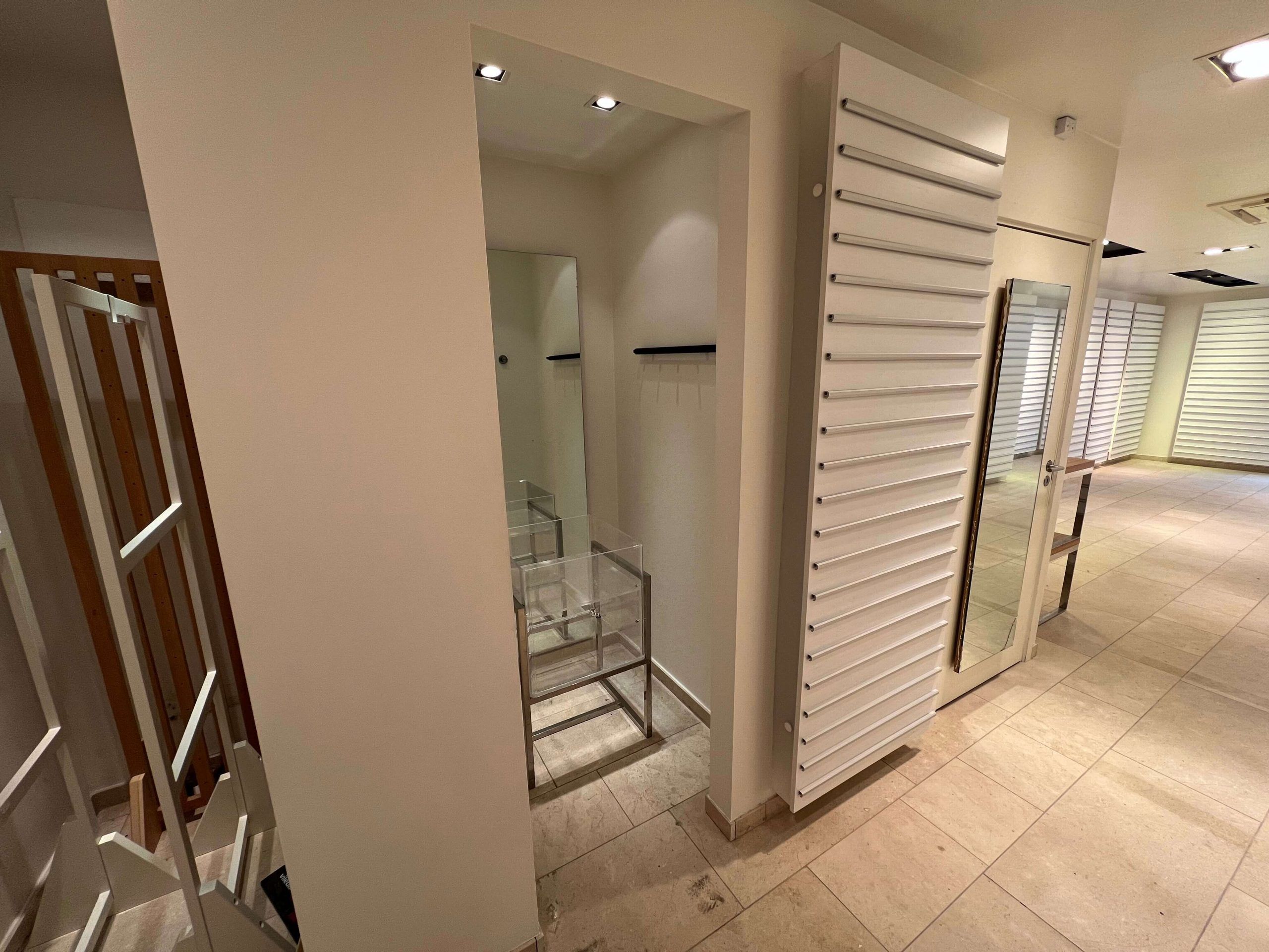 En lukket dør og lyse loftslamper i en indgangsrum, med et spejl og et skab med åbne hylder og en glas stang.