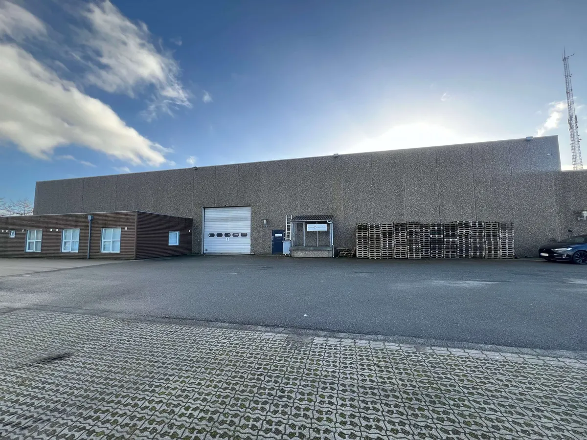 Et stort, industrielt lagerbygning med ståldør og glasvinduer og et parkeringsområde foran med et parkeret bil