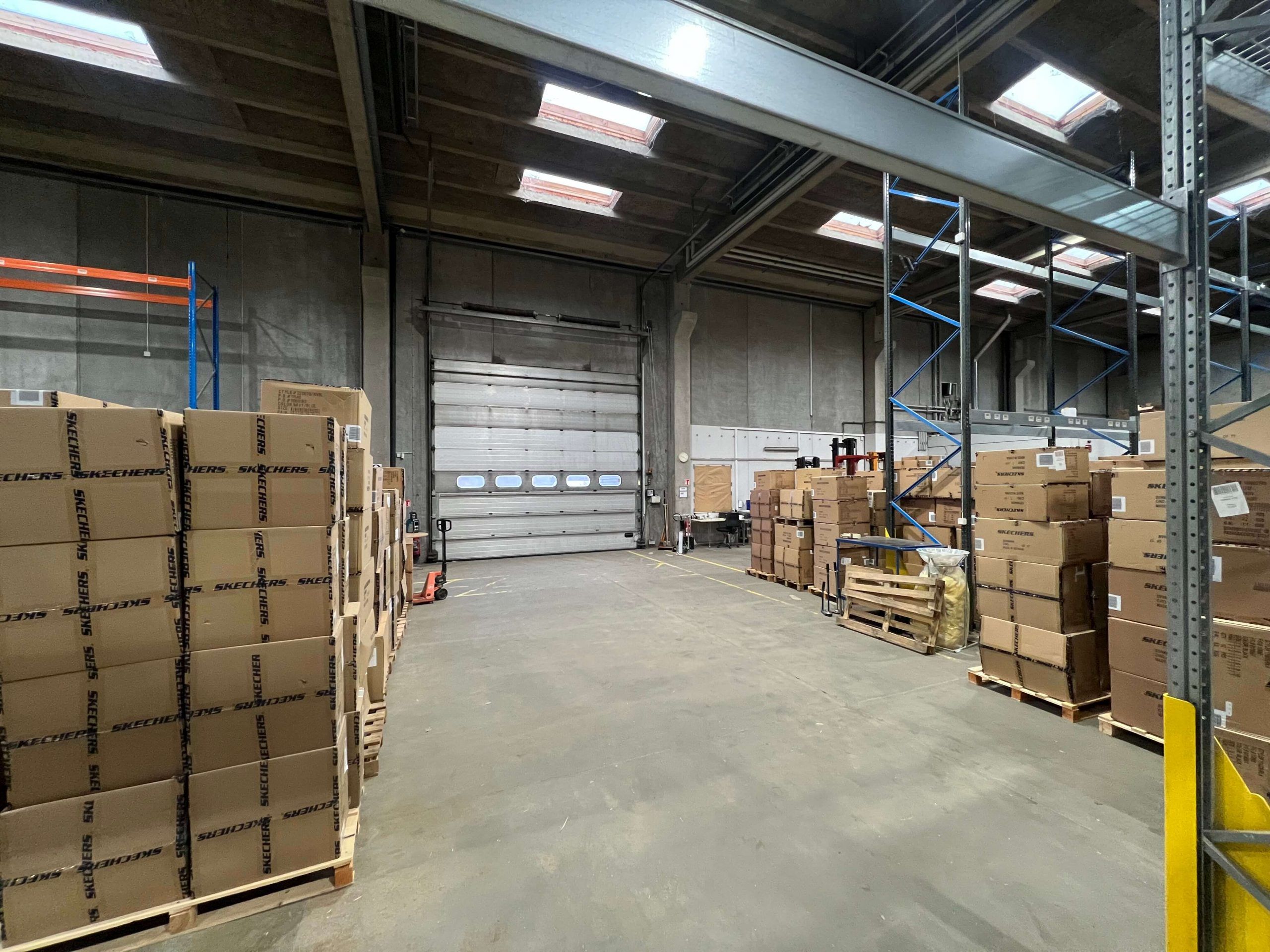 Lager med flere kartoner, der indeholder Skechers. Højt gulv med store åbninger og skyggefuldt lys.