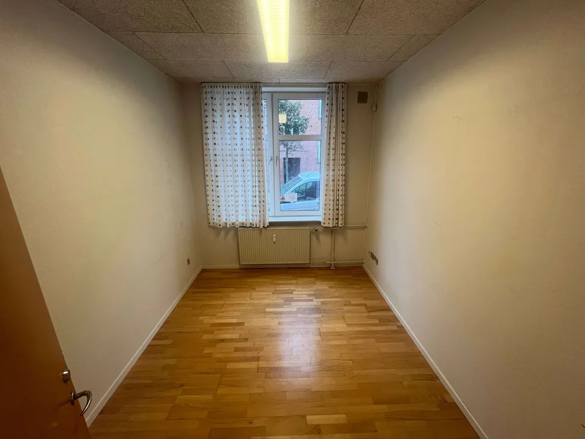 En tom lejlighed med lysegule vægge, trægulv, hvide gardiner og radiator under vinduet med udsigt til en parkeringsplads med