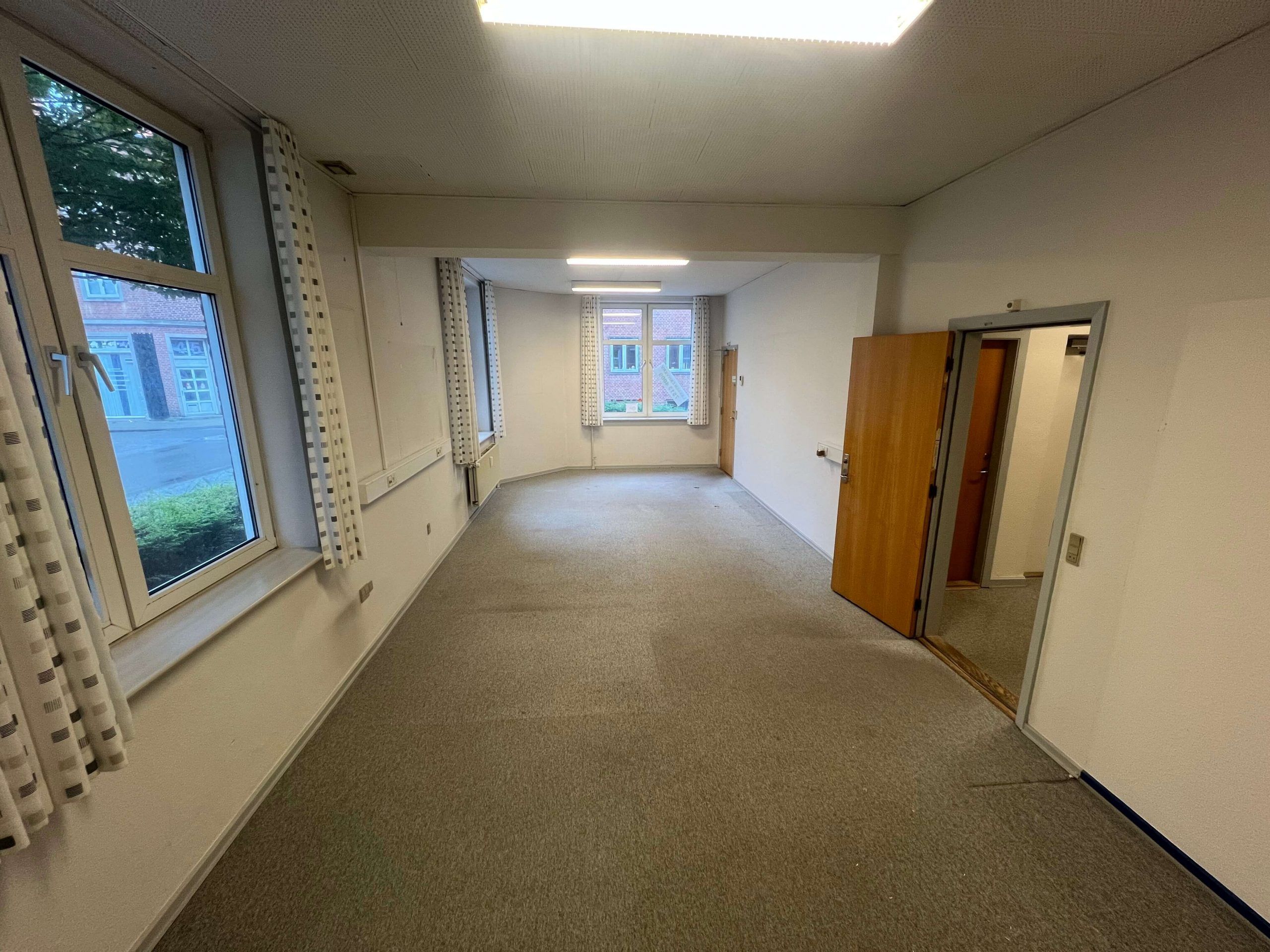 En lang hal med lysegul gulvbelægning og hvide vægge samt fire vinduer med hvide ruder og gardiner