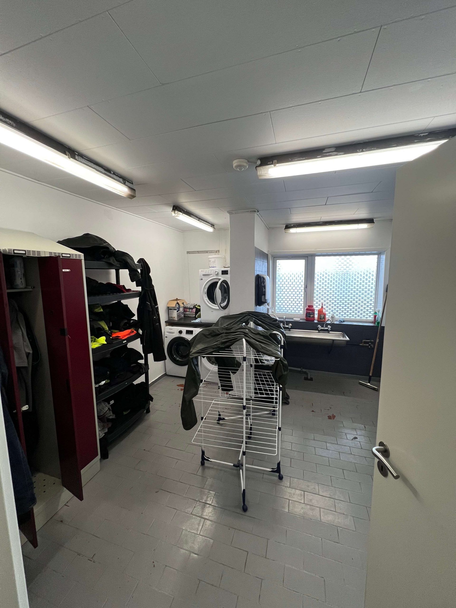 En vaske- og omklædningsrum med en hvid gulv og to vaskemaskiner og tørretumblere, samt et hængestativ og en åben dør