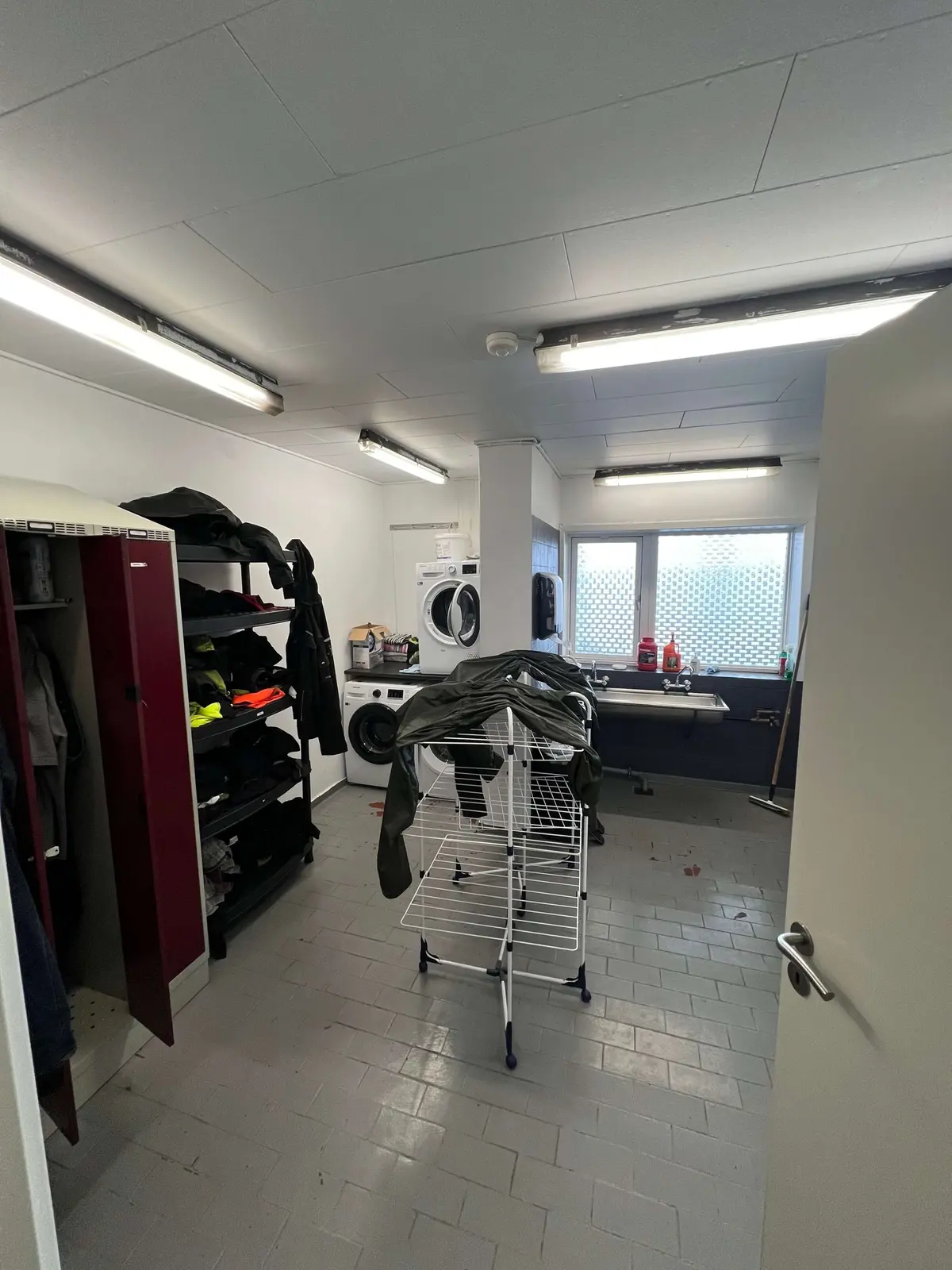 En vaske- og omklædningsrum med en hvid gulv og to vaskemaskiner og tørretumblere, samt et hængestativ og en åben dør