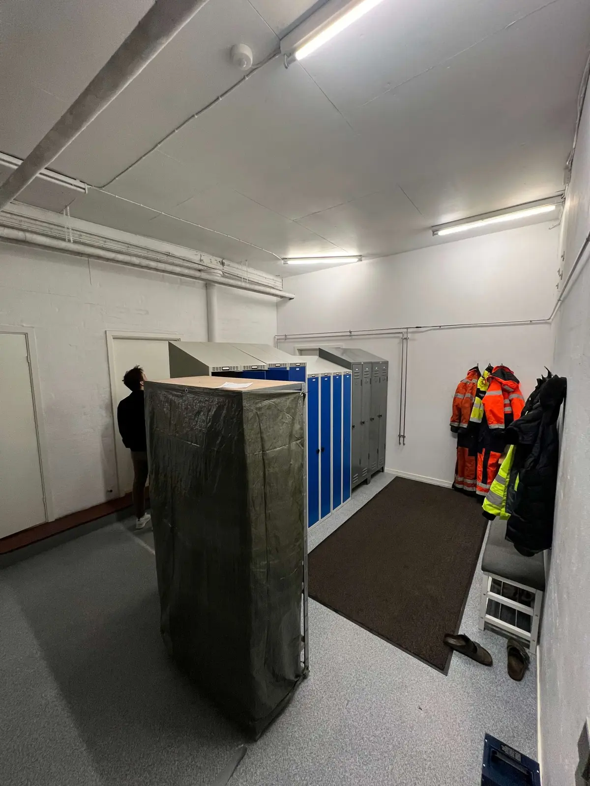 Person står ved indgangen til garderobelokaler med hængende tøj og sko, tørreloft, metal og træelementer, samt gulv og