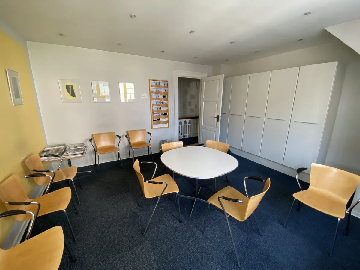 En kontorlokal med hvide vægge og lysegul væg, møblerne er i beige og hvide farver, der er en stor rund bord med flere stole