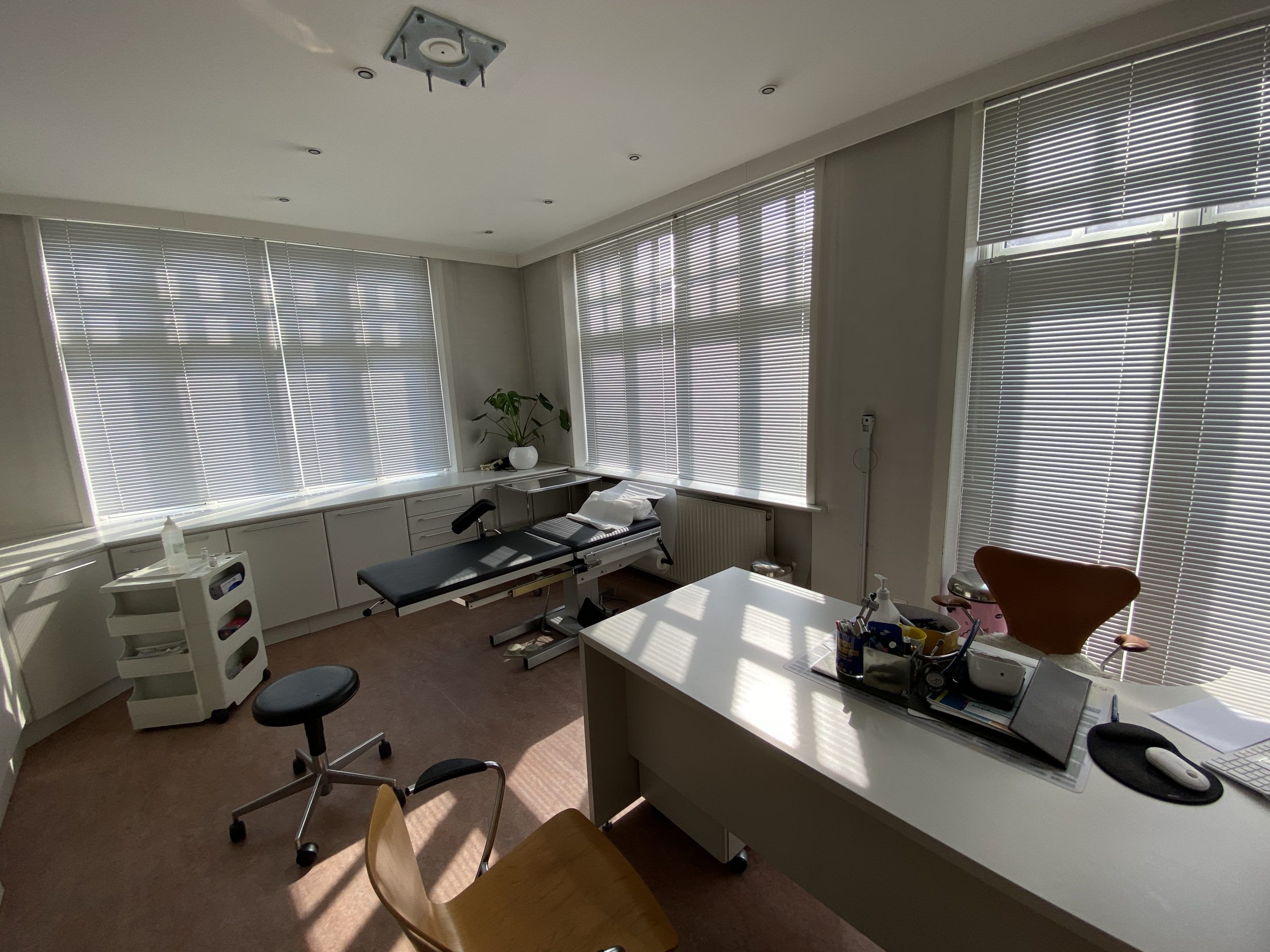 En lægepraksis med en stor, hvid skrivebord, en højstole og en eksaminationsseng med puder og et håndskudstativ
