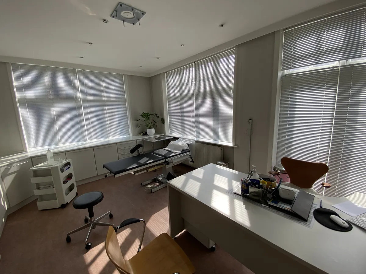 En lægepraksis med en stor, hvid skrivebord, en højstole og en eksaminationsseng med puder og et håndskudstativ