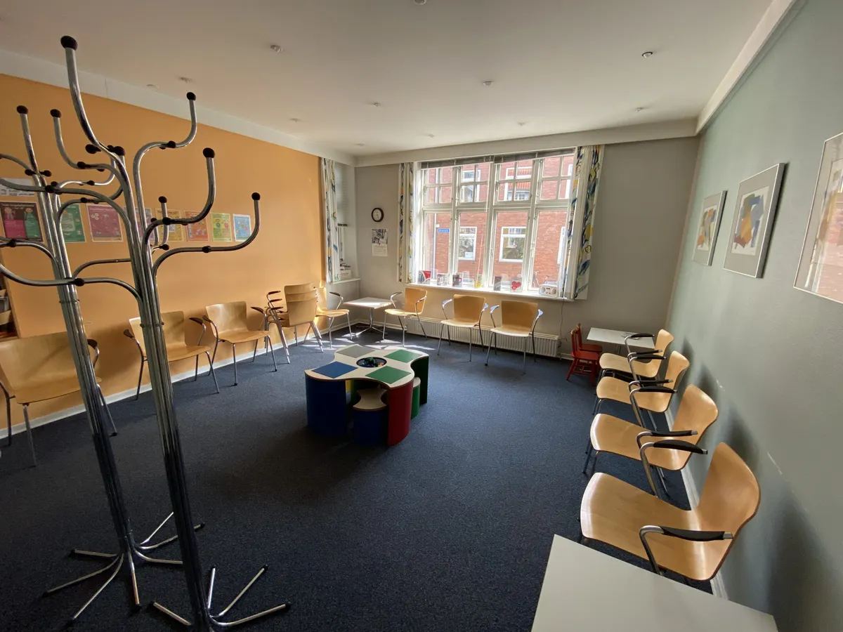 En stor, møbleret stue med en børnehøj stol og et rekreativt område med stole og små borde og vinduer med udsigt til bygning