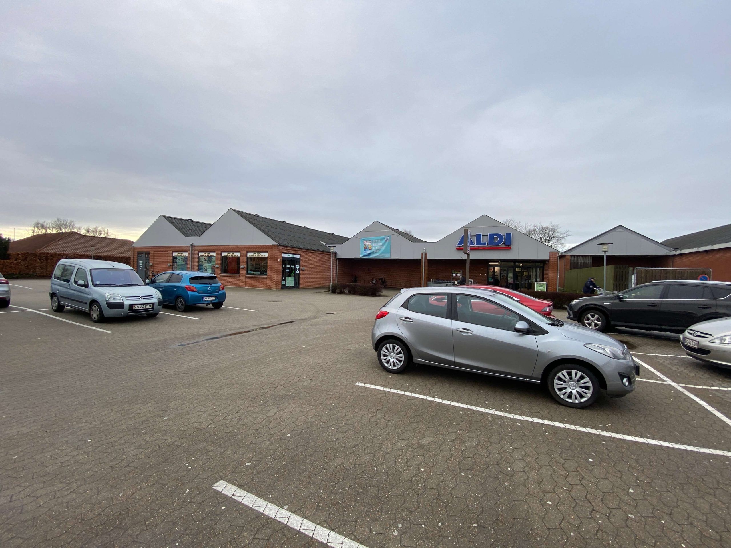 En række forretninger med Aldi-butik i forgrunden og flere parkerede biler i parkeringspladsen