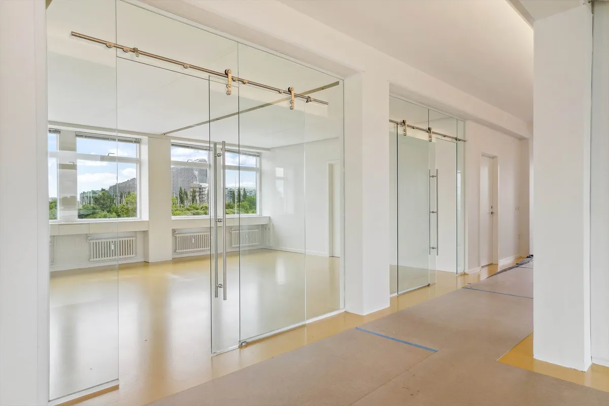 Rummeligt kontor med store glasruder og trægulve i et lyst rum, der har udsigt til byen og grønne træer