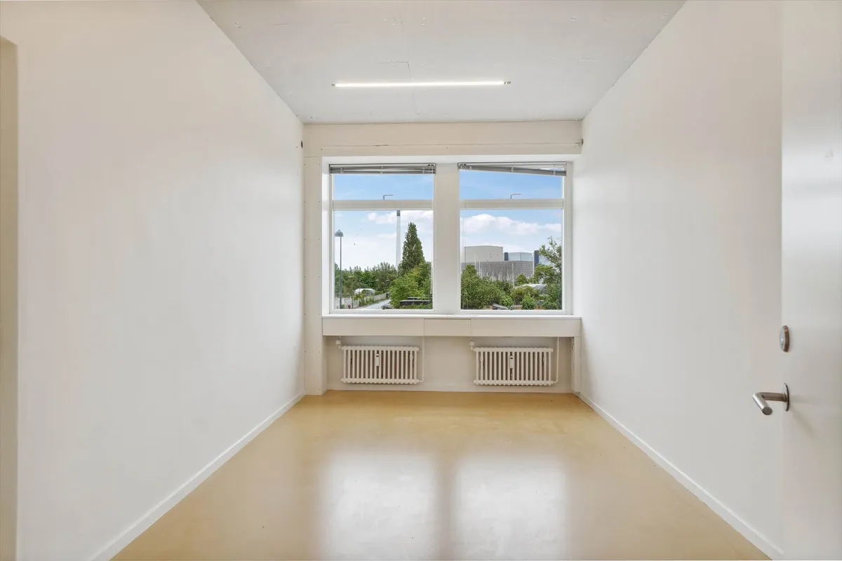 Et lyst kontor med hvid væg og hårdt trægulv samt radiatorer under vinduet og en dør med håndtag