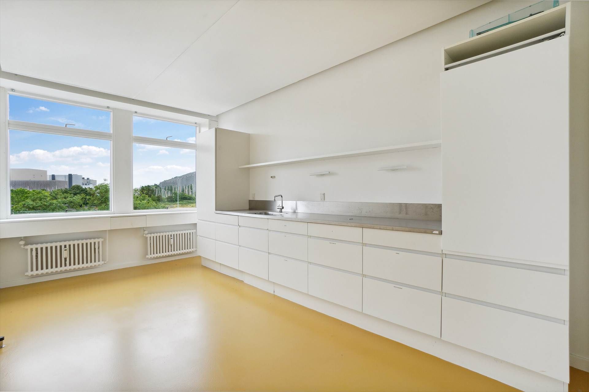 En lys og rummelig køkken med hvide vægge, højloft, gult gulv og lysevinduer med udsigt over bygninger og grønne træer