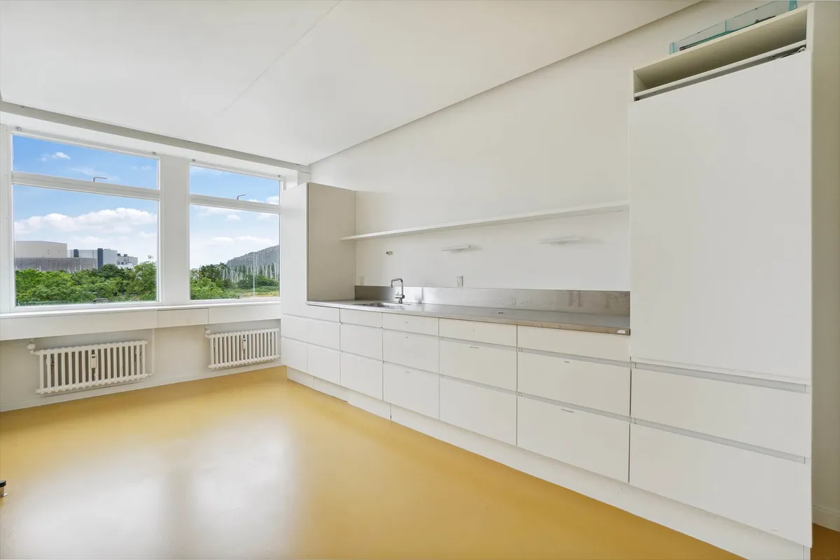 En lys og rummelig køkken med hvide vægge, højloft, gult gulv og lysevinduer med udsigt over bygninger og grønne træer