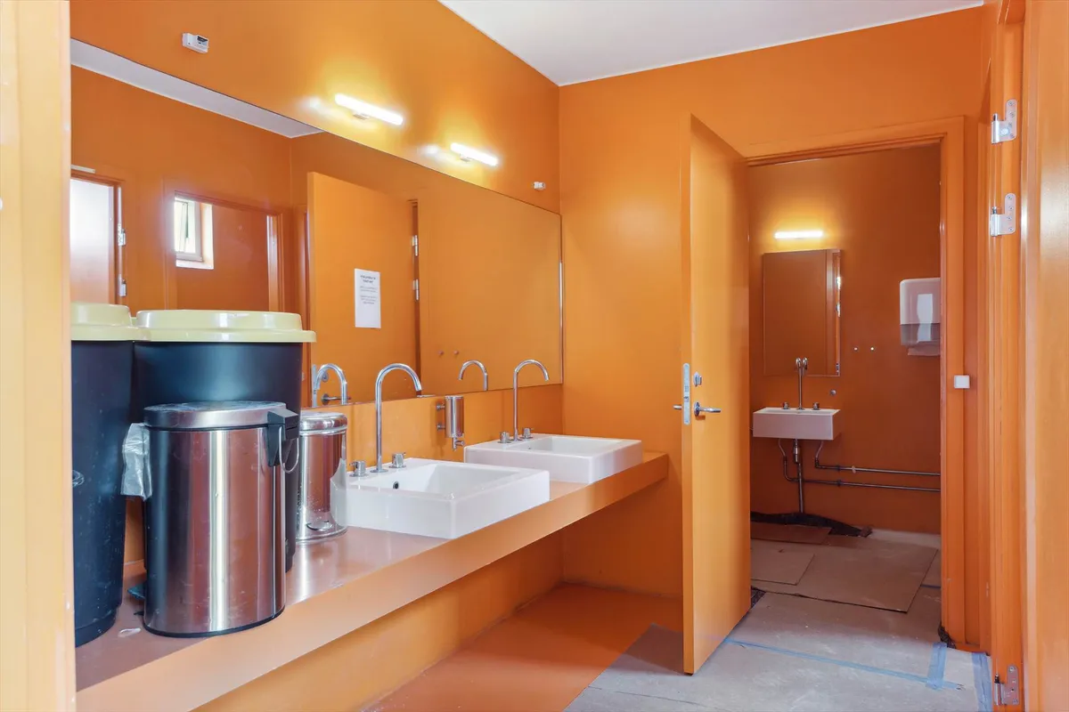 To lyse orange toiletrum med lysegult gulv og to dobbeltvask med håndklædeholdere og højtryksblæsere, samt et skraldespand